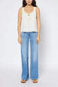 Woman wearing a sleeveless cream top with scalloped edges, blue wide-leg jeans, and sandals, standing against a plain white background.