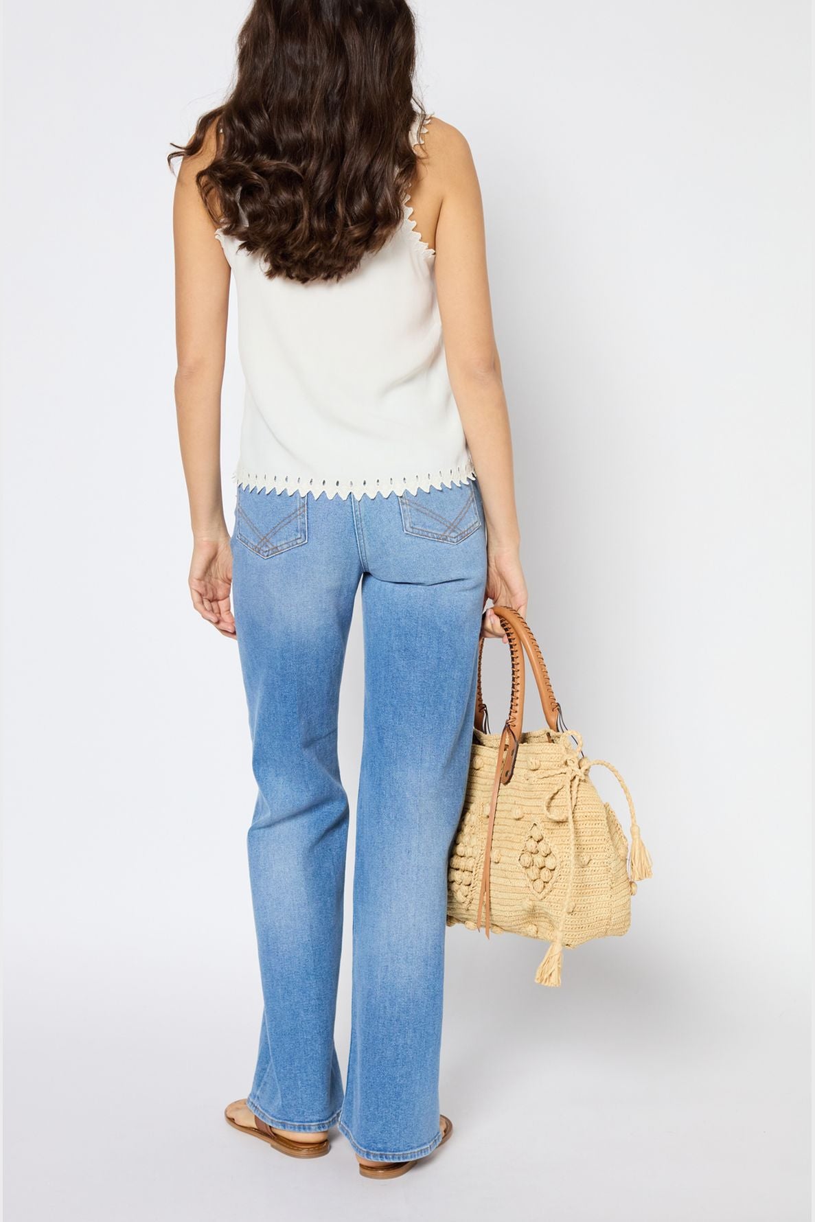 Woman standing facing away, wearing a white sleeveless top, light blue jeans, and sandals, holding a woven straw bag with wooden handles and tassels. Her hair is long and wavy. Background is plain white.