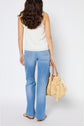 Woman standing facing away, wearing a white sleeveless top, light blue jeans, and sandals, holding a woven straw bag with wooden handles and tassels. Her hair is long and wavy. Background is plain white.