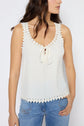 Woman wearing a sleeveless white top with scalloped edges and lace-up front detail, paired with blue jeans, standing against a plain background.