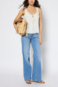 A woman in a sleeveless white top, light blue flare jeans, and brown sandals holds a woven straw bag. She stands against a plain white background.