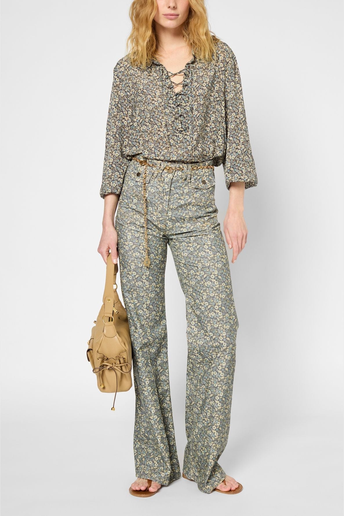 A woman wearing a chemise à fleurs with an encolure lacée and matching high-waisted, flared pants holds a tan handbag. She embodies un style classique chic with her wavy blonde hair and brown sandals.