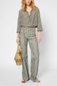 A woman wearing a chemise à fleurs with an encolure lacée and matching high-waisted, flared pants holds a tan handbag. She embodies un style classique chic with her wavy blonde hair and brown sandals.