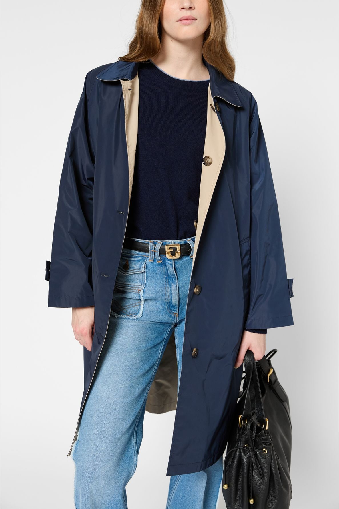 Woman wearing a navy blue trench coat over a dark sweater and blue jeans, holding a black handbag. Only her head and upper body are visible against a plain background.
