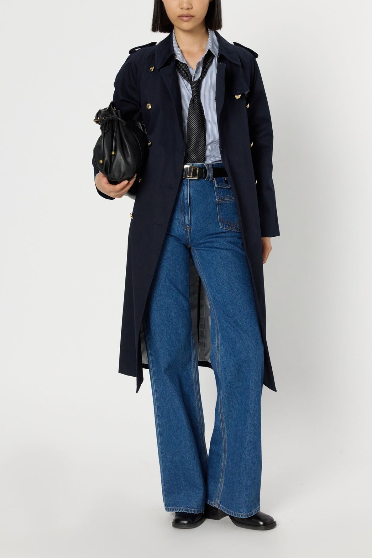 A person wearing a dark trench long coat with boutons dorés, a blue shirt, black tie, wide-leg blue jeans, and black shoes holds a black bag. Their head is partially out of frame against a white background.