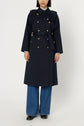 A person wearing a manteau femme trench long navy coat with boutons dorés, blue jeans, and black shoes stands against a plain white background.
