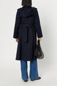 Person standing with their back facing the camera, wearing a dark blue trench long manteau femme with boutons dorés, paired with blue jeans and black shoes, holding a black handbag in their left hand against a plain background.