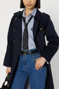Woman wearing a navy trench long manteau femme with elegant boutons dorés, a striped button-up shirt, polka dot tie, blue jeans, and holding a black bag. Her hair is straight and chin-length.