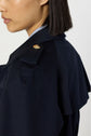 A close-up side view of a person wearing a dark navy manteau femme trench long with a shoulder epaulet and boutons dorés, showing a short bob haircut and part of a collared shirt underneath.