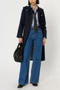 A person wearing a long black trench coat with boutons dorés, a blue shirt, black tie, high-waisted wide-leg blue jeans, and black shoes, holding a black handbag. Their face is partially visible.