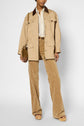 A person wearing a tan utility jacket in coton with a col velours côtelé, beige shirt, patterned tan pants, tan heels, and carrying a small brown shoulder bag, posing against a plain background.