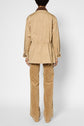 A person stands facing away, wearing a light tan trench mi-long with an elastic waist and tan patterned pants. The jacket appears to be crafted from coton, adding a touch of understated elegance. The background is plain white.