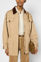 A person wears a beige col velours côtelé jacket with a brown collar, striped shirt, tan patterned pants, and carries a brown suede handbag with a gold chain strap. Only the lower face and torso are visible.