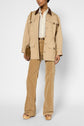 Person wearing a beige col velours côtelé jacket with brown collar, cream shirt, tan patterned pants, platform sandals, holding a brown shoulder bag, standing against a plain white background.