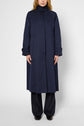 A person wearing a long imperméable bleu marine with black buttons, black pants, and black shoes stands against a plain white background. Their face is partially out of frame.