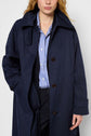 A person wearing an imperméable bleu marine Palto over a blue and white striped shirt, posing with one hand in their pocket against a plain light background.