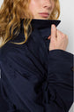 A person with wavy, light brown hair wears an imperméable bleu marine, holding the collar up close to their face. Only the lower part of their face and hand are visible.