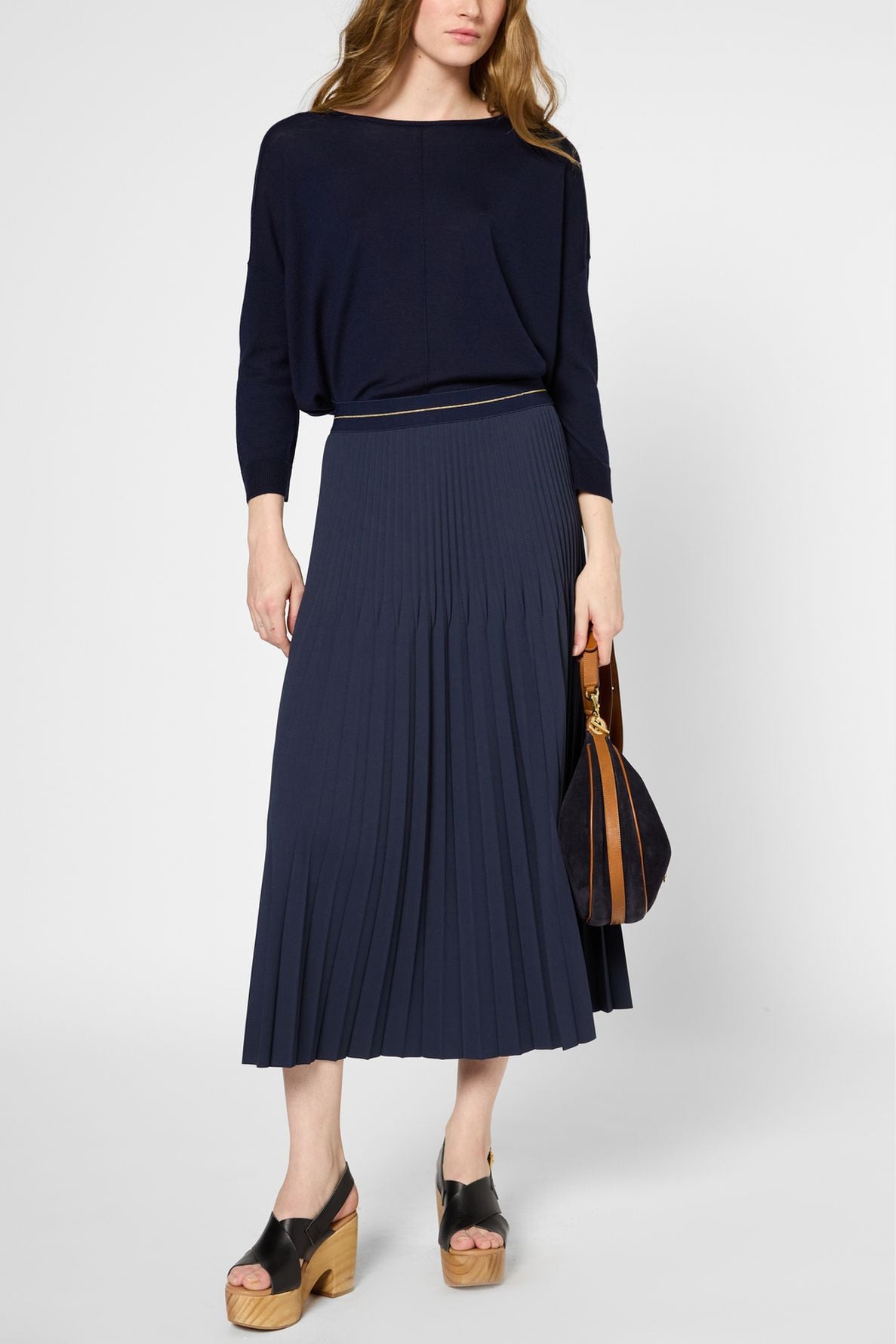 A woman wearing a loose navy top with a matching pleated midi skirt, holding a brown handbag, and wearing platform sandals.