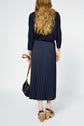 A woman with long wavy hair, dressed in a navy top and pleated navy midi skirt, stands facing away while holding a black purse. She wears black sandals with chunky wooden heels.