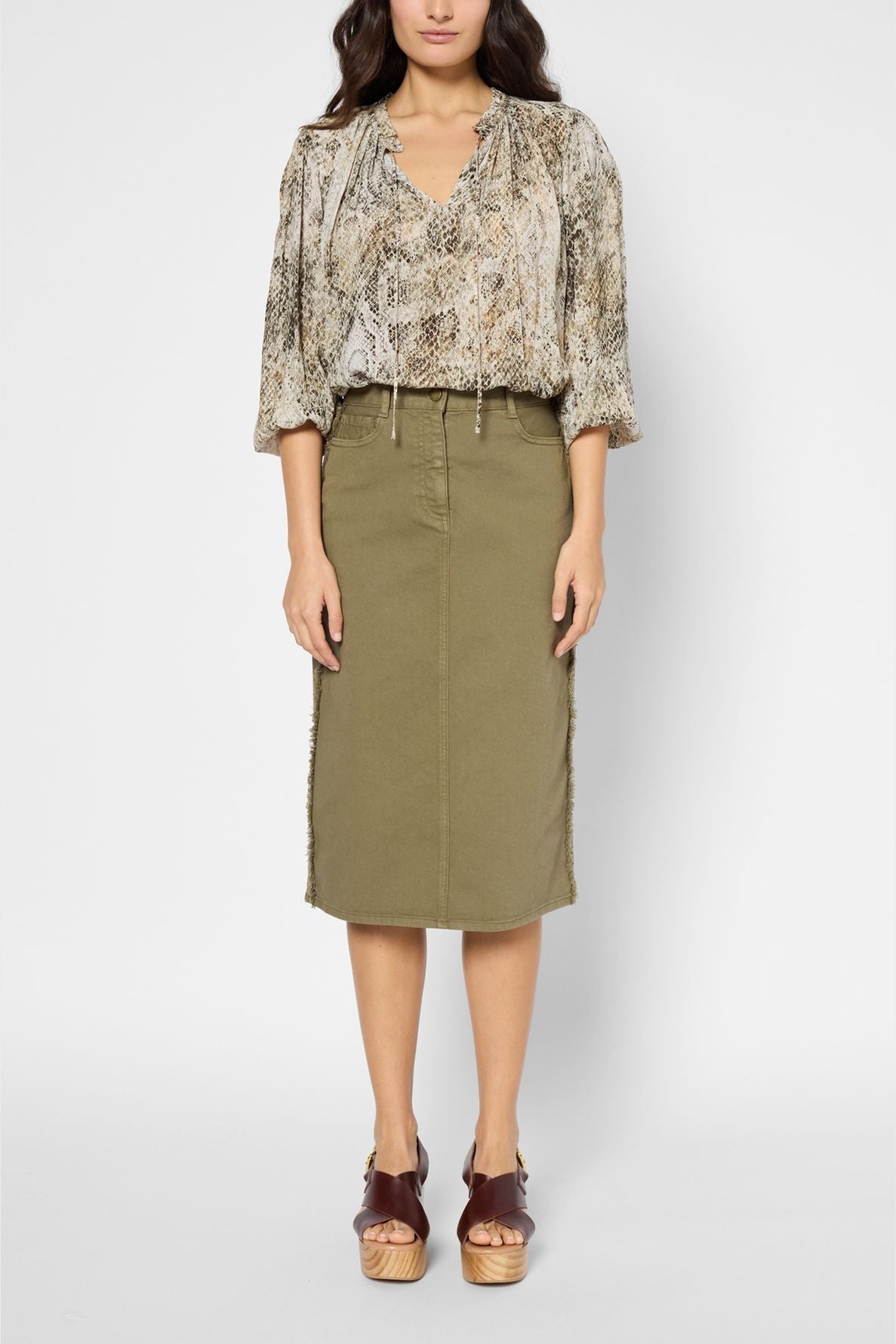 Woman wearing a snakeskin-print blouse, olive green knee-length skirt with frayed sides, and brown platform sandals, standing against a plain white background.