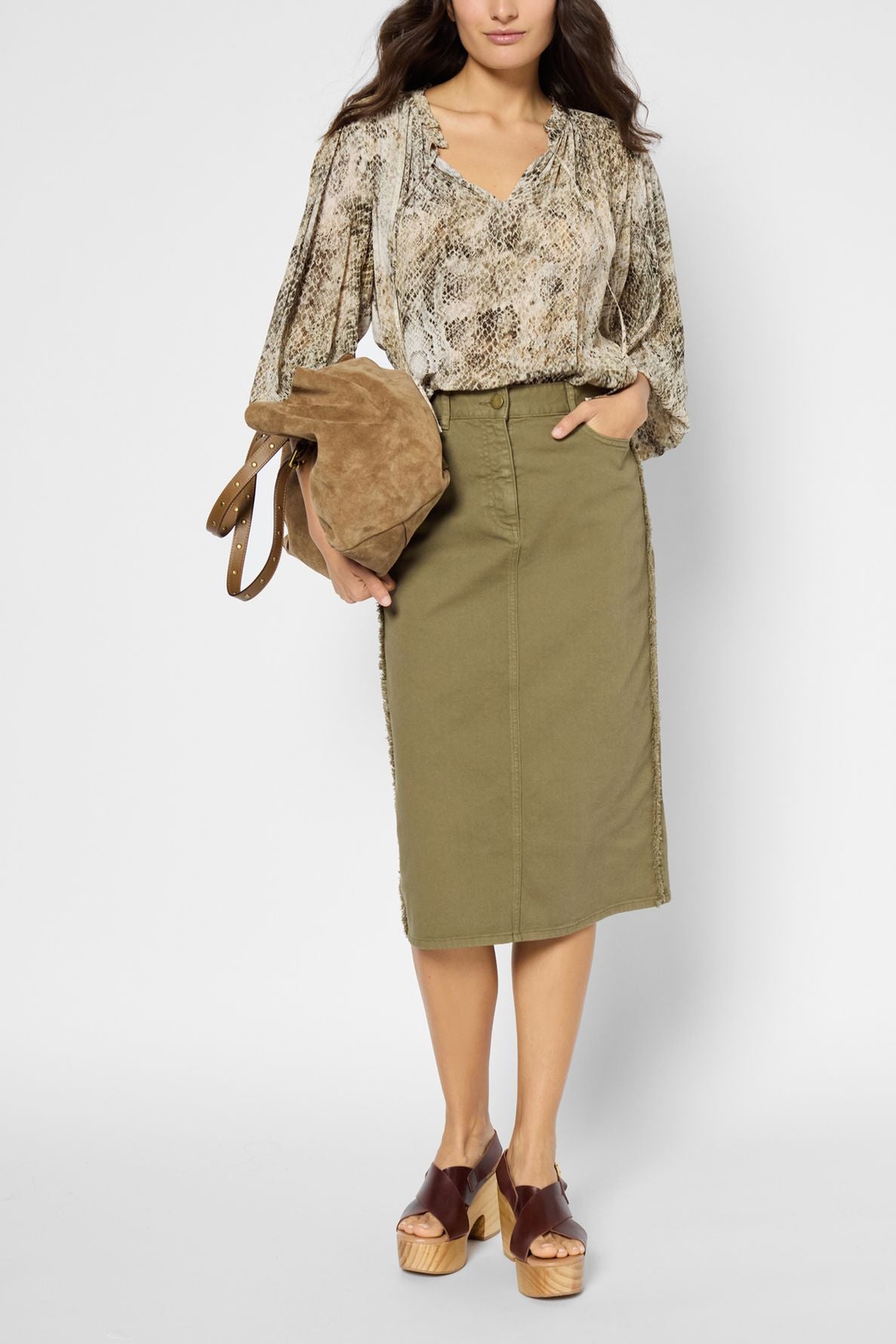 Woman wearing a snakeskin-patterned blouse, olive-green midi skirt, brown platform sandals, and holding a tan suede bag, standing against a plain white background.