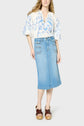 A woman wears a white blouse with blue floral embroidery, a light blue denim midi skirt, and brown platform sandals. She stands against a plain light gray background.