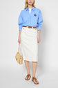 A woman wears a light blue button-up shirt, a jupe en denim blanc mi-longue, brown sandals, and carries a woven handbag. She also has a brown belt and her hair is loose. The background is plain and light-colored.