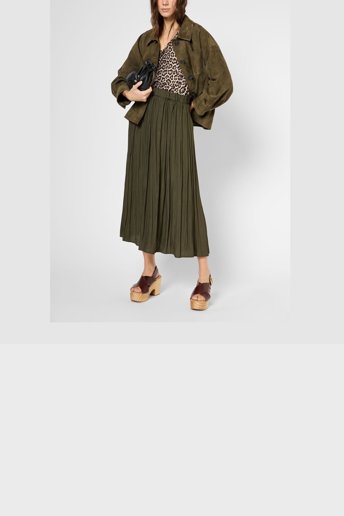 A woman wearing a brown suede jacket, leopard print top, olive green jupe plissée midi skirt, and wooden platform sandals, holding a black clutch.