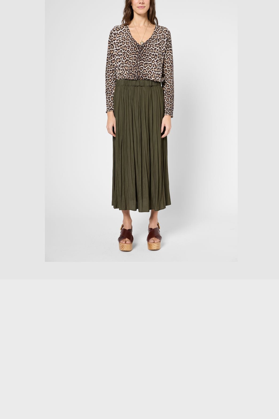 A woman stands facing forward, wearing a long-sleeve leopard print blouse, an olive green jupe plissée midi skirt, and brown platform sandals. The background is plain white.
