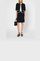 Woman wearing a jupe tailleur in coton mélangé with white trim, featuring a short-sleeved jacket and black sandals, holding a black handbag. She stands against a plain background. Her face is partially obscured by her hair.
