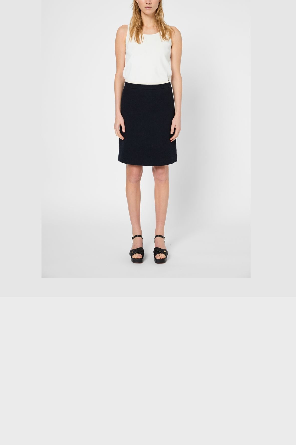 A woman stands facing forward in a sleeveless white top crafted from coton mélangé, a black jupe courte, and black sandals against a plain white background. Her arms are relaxed by her sides.