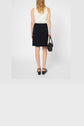 A woman with blonde hair, wearing a sleeveless white top made from coton mélangé, a black jupe courte, and black sandals, stands facing away and holds a black handbag at her side against a plain white background.