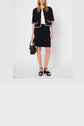 A woman wearing a jupe tailleur noire and a matching short-sleeve jacket with white trim, crafted from coton mélangé, holds a black handbag and wears black sandals as she stands against a plain background.