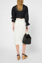 Woman standing with her back to the camera, wearing a black blouse and a jupe droite blanche, paired with black platform sandals and holding a black handbag with a gold chain handle.