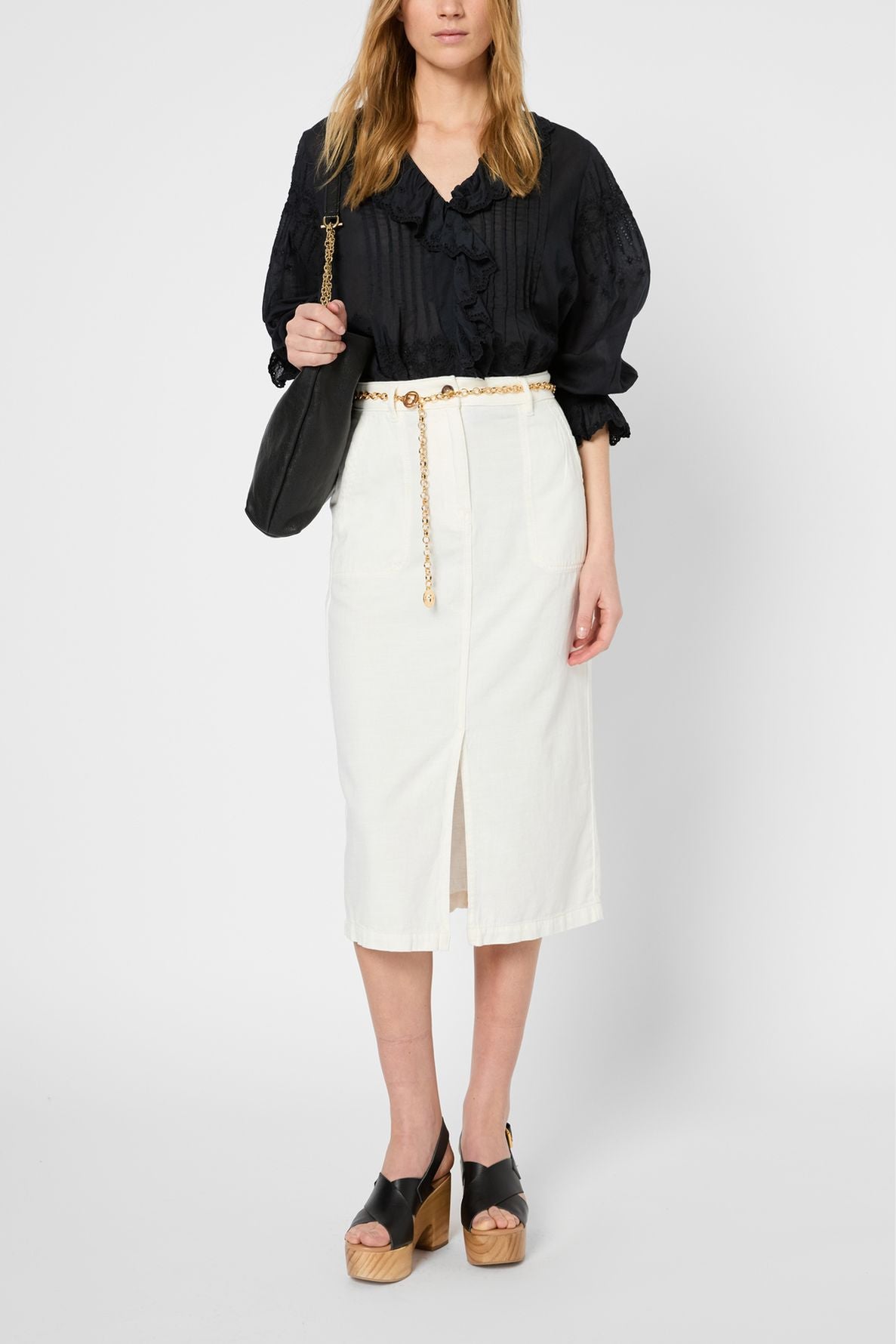 Woman wearing a black blouse, a jupe taille haute — a white jupe droite blanche mi-longue with a front slit, black wedge sandals, and carrying a black shoulder bag, standing against a plain light background.