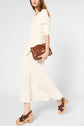 A woman in a cream ribbed sweater and matching skirt walks while holding a brown leather clutch. She wears brown platform sandals with wooden heels. The background is plain white.