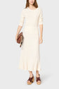 A woman wearing a long cream ribbed knit dress, brown platform sandals, and holding a brown quilted clutch bag, standing against a plain light background.
