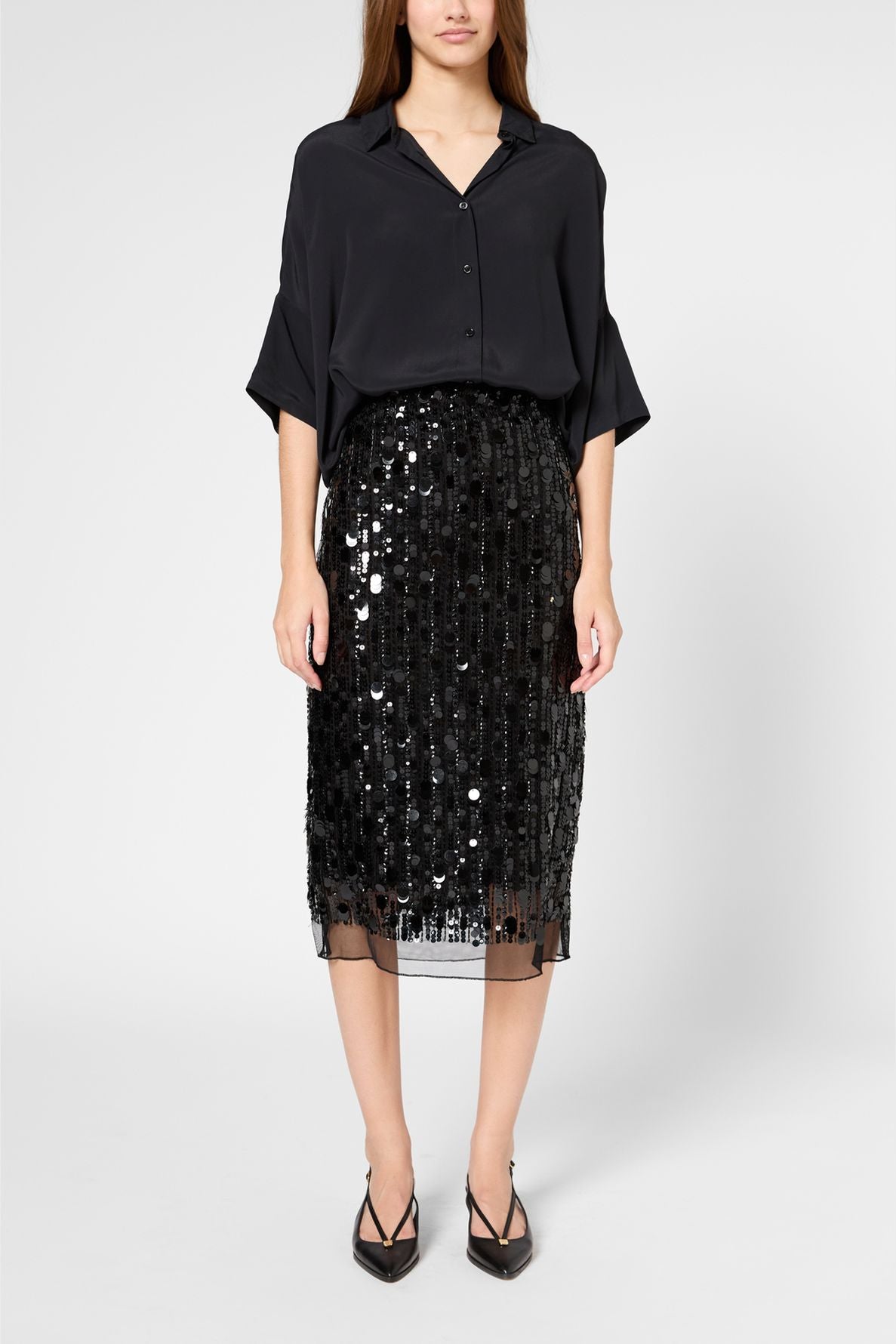 A woman wearing a loose black blouse, a black sequin jupe droite longueur midi, and black pointed heels stands against a plain light background.