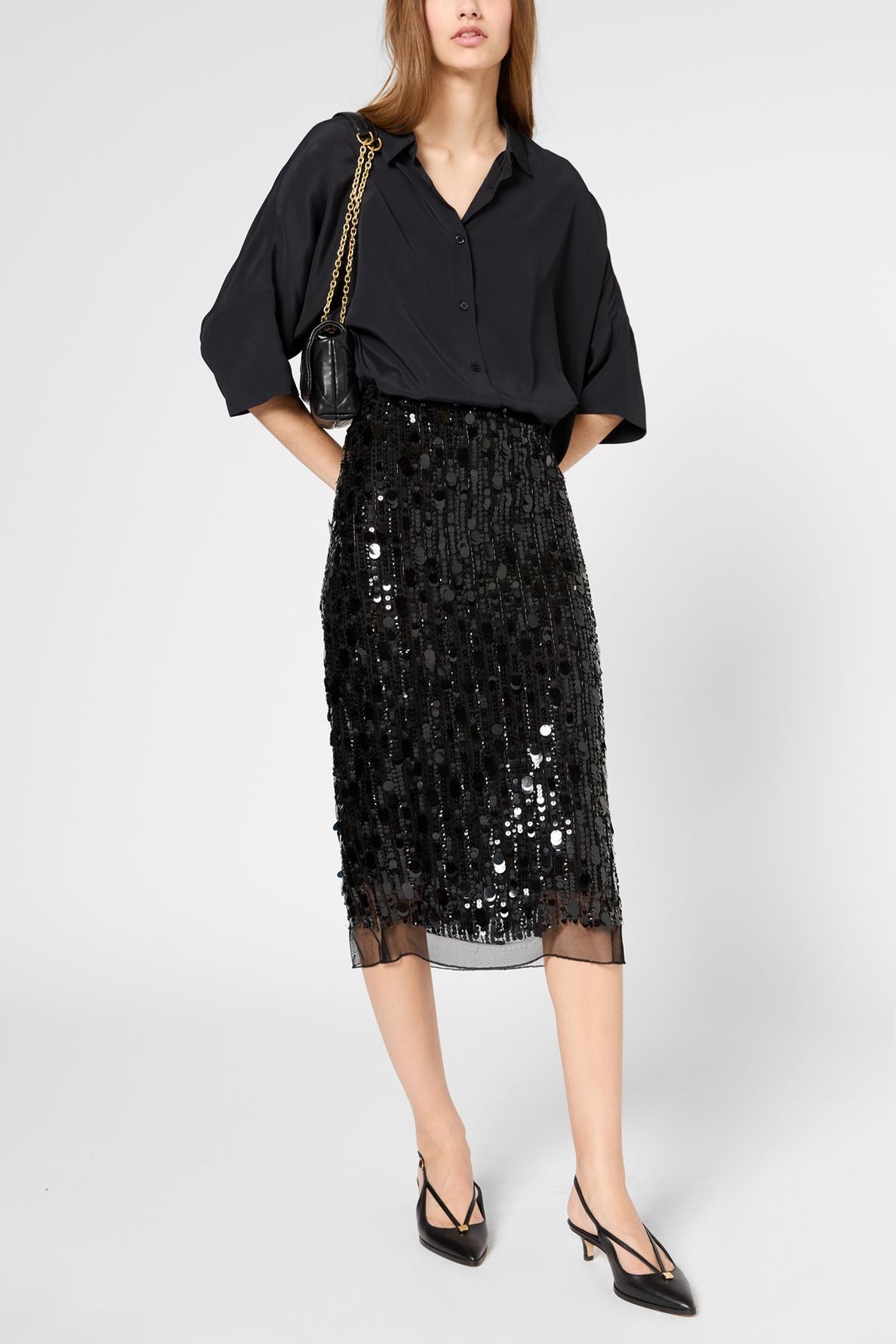 Woman wearing a loose black button-up shirt, a black jupe droite à sequins longueur midi, black slingback heels, and carrying a small black purse with a gold chain. She stands on a plain light gray background.
