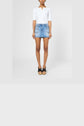 A person wearing a white short-sleeve top, a blue mini-jupe en denim from the collection Studio, and black sandals stands against a plain light background.