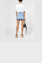 Person standing with back to camera, wearing a white shirt and a mini-jupe en denim bleu clair de la collection Studio, paired with wooden platform sandals and holding a black handbag against a plain background.