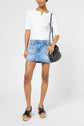 A person wearing a white short-sleeve top, une mini-jupe en denim from the collection Studio, black platform sandals, and holding a black handbag, standing against a plain light background.