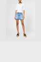A woman stands wearing a white short-sleeve top, a mini-jupe en denim bleu clair avec des poches à l’avant, and black platform sandals. The look is part of the collection Studio, set against a plain, light-colored background.