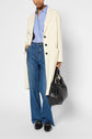 A person wearing a cream manteau laine over a blue striped shirt, blue wide-leg jeans, and black loafers holds a large black handbag.