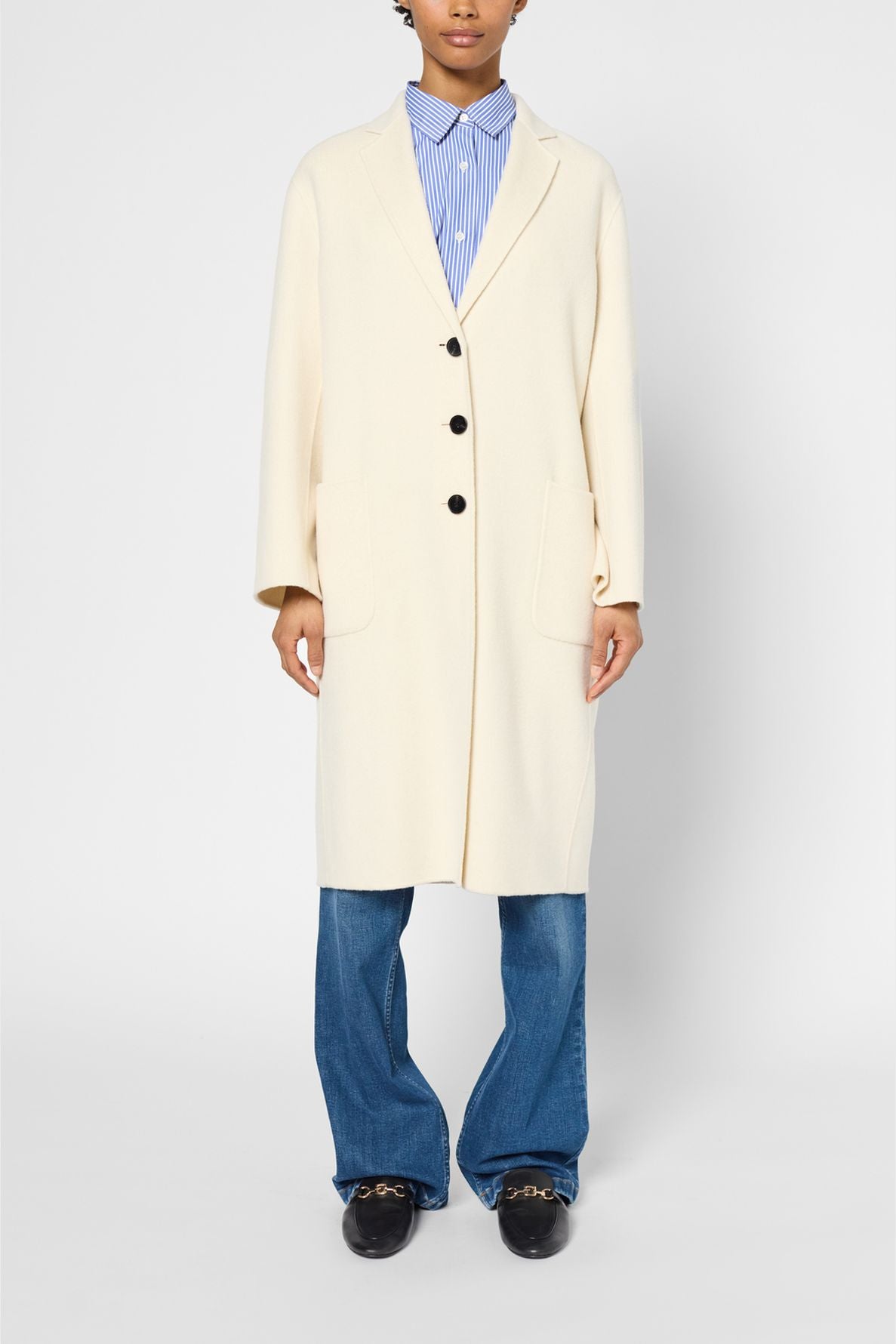 Person wearing a manteau mi-long cream-colored coat with black buttons over a blue striped shirt, blue jeans, and black loafers, standing against a plain light background.