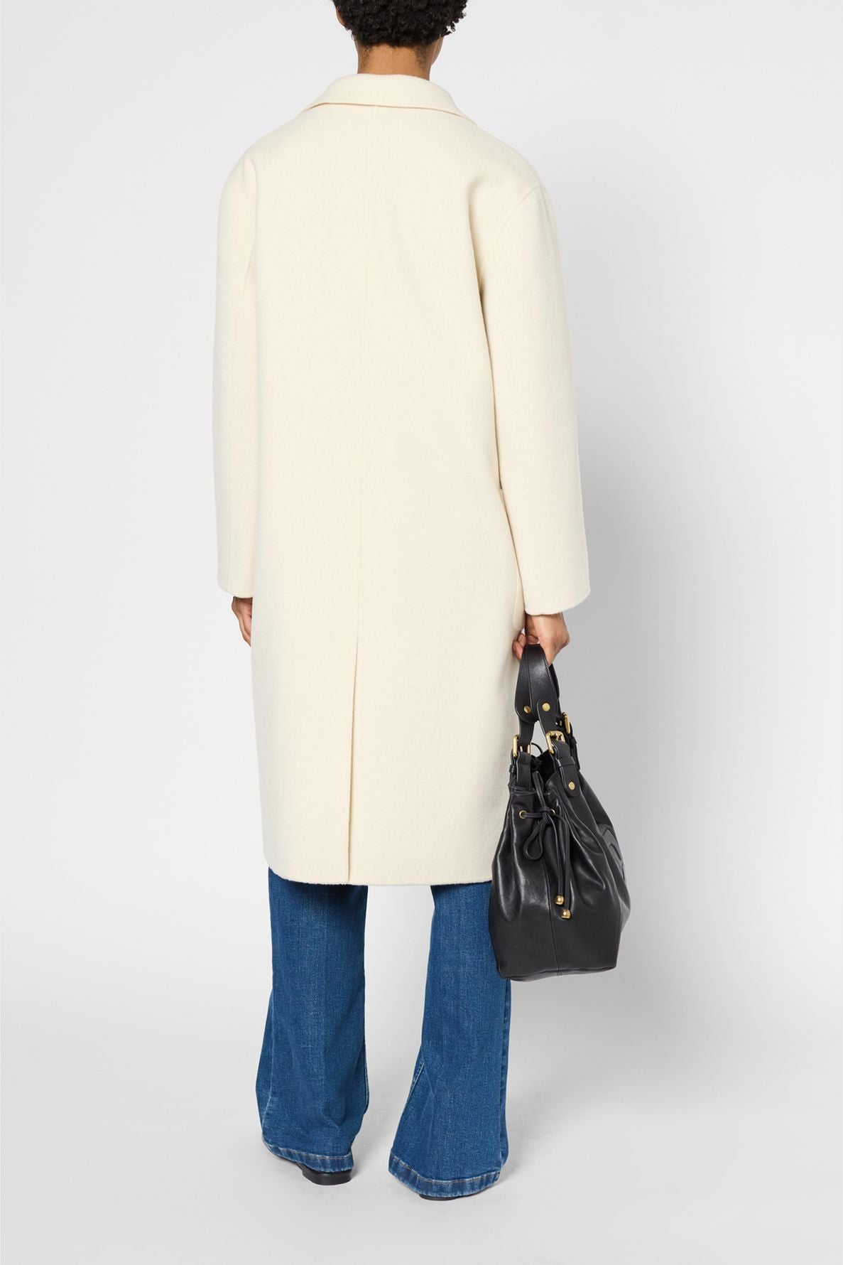Person facing away, wearing a manteau mi-long cream-colored coat over blue jeans, holding a large black handbag in their right hand. The background is plain and light-colored.