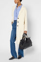 A person wearing a manteau droit femme in cream over a blue striped shirt and blue jeans, holding a large black handbag, and wearing black loafers, poses against a plain background.