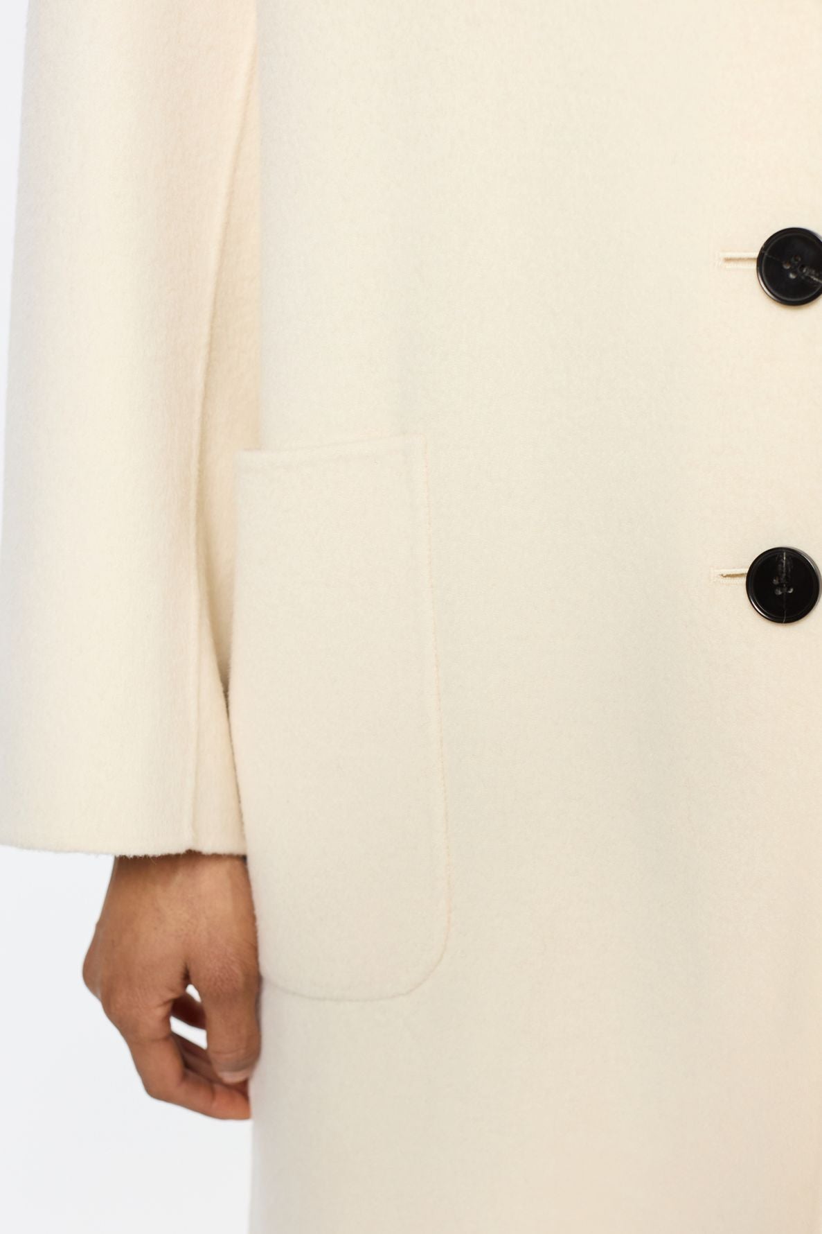 A close-up of a person wearing a cream-colored manteau droit femme with black buttons, showing one sleeve, a front pocket, and a hand partially visible by the pocket.