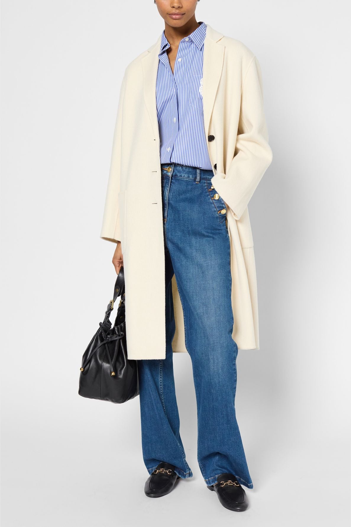 Person wearing a manteau droit femme in cream—a long, tailored coat—over a blue striped shirt, blue jeans, and black loafers, holding a black handbag. Face is partially out of frame. Background is plain white.