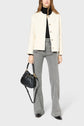 A woman wearing a veste rétro chic cream jacket, black turtleneck, gray flared jeans, and black shoes holds a black handbag. She stands against a plain light gray background.